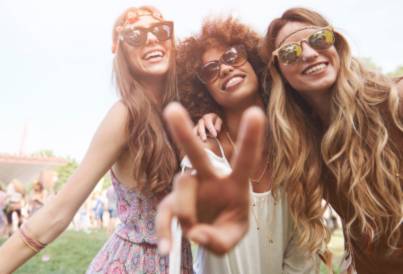 Three women with long hair and sunglasses posing outdoors, one making a peace sign.